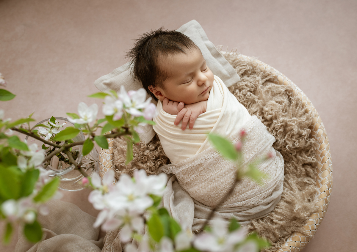 Schlafendes Neugeborenes, eingewickelt in helle Cremetöne, liegt in einem Korb auf weichen Fellen; im Vordergrund blühen zarte weiße Frühlingszweige.